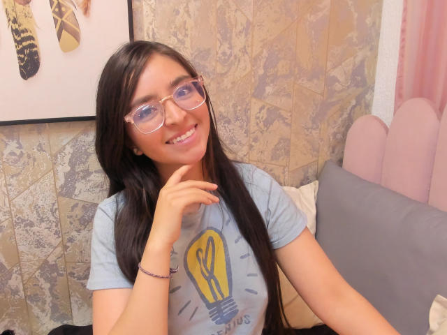 EvaMcAllister with long dark hair, glasses, smiling, light blue shirt with yellow light bulb, pink wrist bracelet, hand near mouth, sitting on gray bed, beige wallpaper.