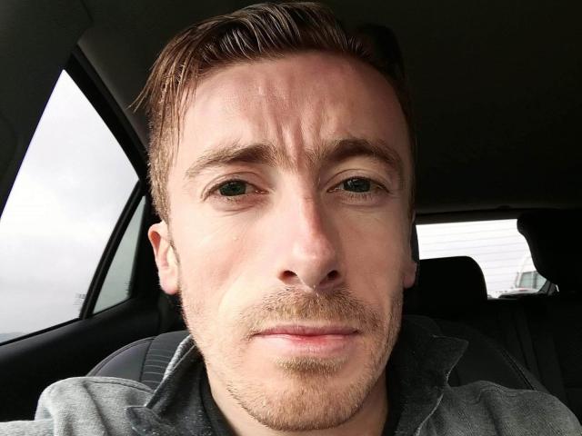 RaviereEight has a serious face, light brown hair, green eyes, and a light beard in a car with a gray jacket. His nose is slightly upturned.