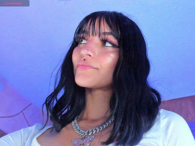 LindaWatsong with black wavy hair, nose ring, and pink eyeshadow, looking up with a slight smile, wearing a white top and a chunky silver necklace with a butterfly pendant.