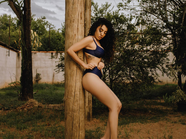 NadiaAdam leans against a wooden pole, wearing navy blue bikini, showing off her tits and ass, with curly black hair, and a tattoo on her right side.