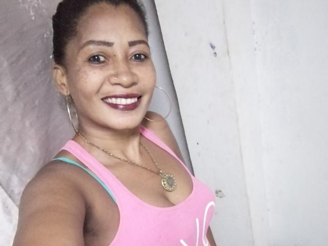 ArnnahZ with brown skin, dark curly hair, hoop earrings, pink tank top showing her boobs, smiling, nose ring, gold necklace, and purple lipstick.