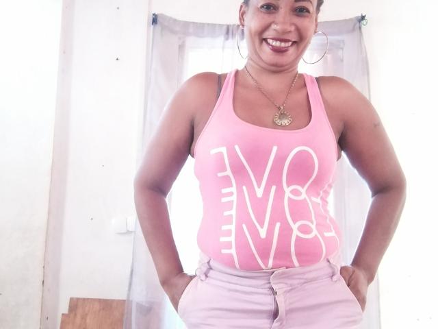 ArnnahZ has medium brown skin, dark hair in a bun, wearing a pink tank top showing her tits, pink pants, and large hoop earrings, smiling with hands on her hips.