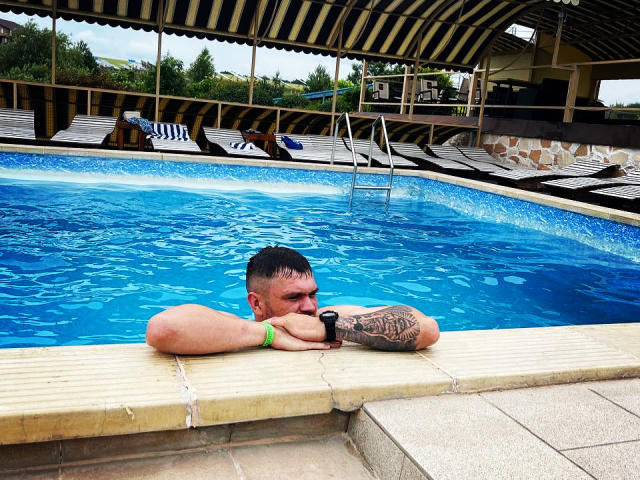 GordonDesire with a tattooed arm, green wristband, and sunglasses, leaning on the pool edge, blue water, striped roof, and wooden loungers in the background.