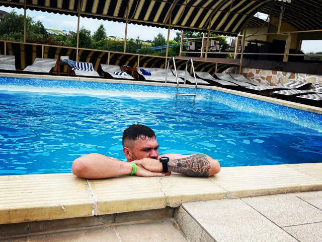 GordonDesire is in a blue pool, arms resting on the edge, wearing a green wristband, tattooed on the right arm, with short dark hair and sunglasses.