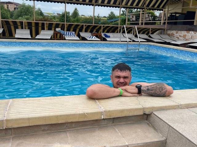 GordonDesire's chubby face with tattoos, green wristband, and black wristwatch, smiling at the camera while arms resting on the pool edge. Blue water in the foreground.