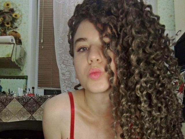 SelenaXCute with long curly brown hair, red bra straps, puffy pink lips, looking slightly to the side, in a cozy room. Her tits are barely visible, showing cleavage.