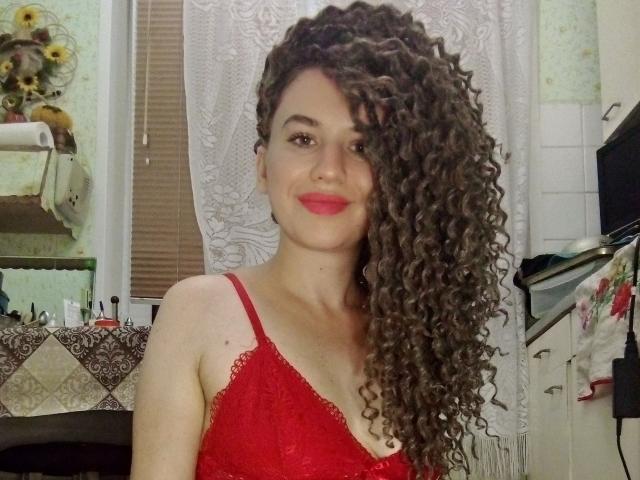 SelenaXCute with long curly brown hair, red lace bra, smiling, showing off her tits, red lipstick, in a bathroom. Her ass is not visible in this photo.