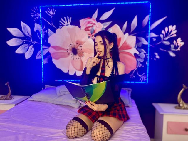 AriannaGray sits on a bed, eating a lollipop, wearing a black off-shoulder top, red plaid skirt, fishnet stockings, and thigh-highs, holding a green notebook.