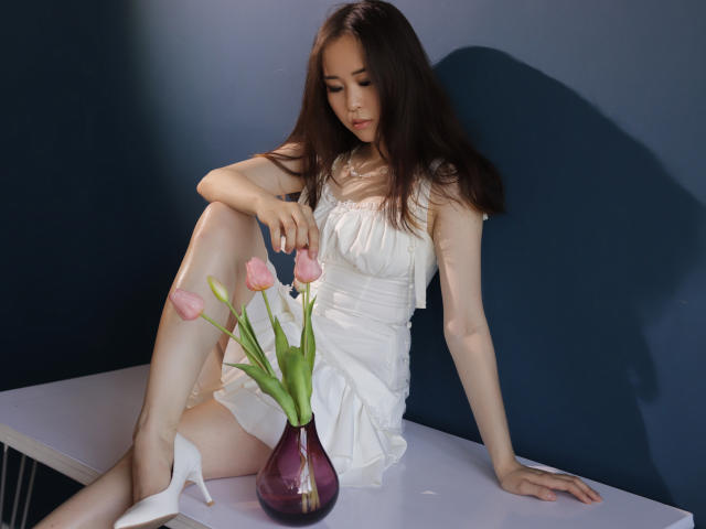 GinnyBean with long brown hair, white dress, and white heels, sits on a table, touching pink tulips in a purple vase, her leg raised, showing her bare ass.
