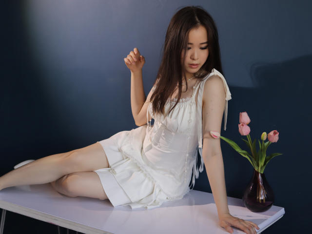 GinnyBean with long brown hair, white dress, and pale skin, sitting on a table, legs crossed, hand raised, pink tulips in a vase on the right.
