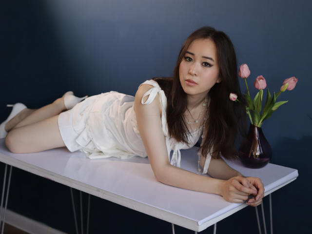 GinnyBean with long brown hair, white dress, and white heels, lying on a table, looking at the camera, with pink tulips in a vase to the right.