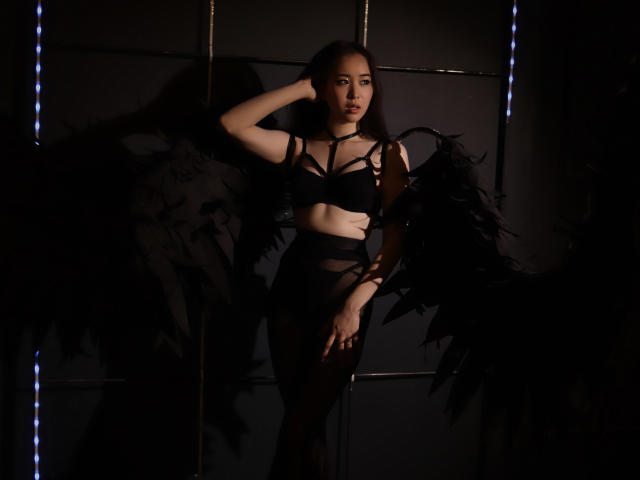 GinnyBean met donkere vleugels, zwarte, open lingerie en bloot achterwerk, staand in de schaduw, hand in haar, verleidelijk naar de camera kijkend, verlicht door blauwe LED-strips..