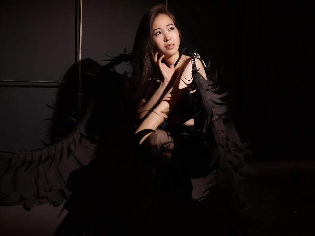 GinnyBean with long brown hair, wearing a black feathered dress, sits in shadow, hand on cheek, soft light on her face, looking slightly away with a gentle expression.