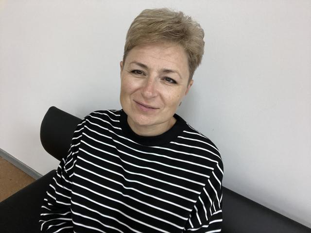 EmiliaGree with short blonde hair, smiling, wearing a black and white striped shirt, sitting on a black chair, has a small nose and slightly wrinkled skin on her face.
