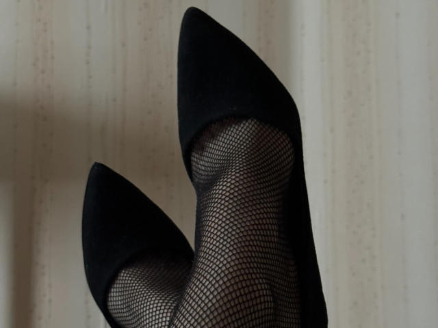 Los tacones negros puntiagudos de Keiri y sus piernas cubiertas con media de red, mostrando sus pies cruzados en el tobillo. Sus dedos apuntan hacia arriba, mostrando su elegante calzado..