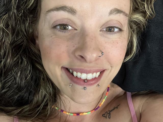 Het gezicht van Harley Quinnzel, met krullend blond haar, groene ogen, een neusring, lip- en kinpiercings, een kleurrijke kralenketting, een roze tanktop en een kleine tatoeage onder de kin..