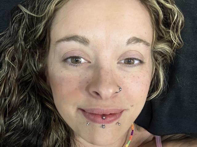 Le visage de Harley Quinnzel, avec ses cheveux blonds bouclés, ses yeux marrons, ses piercings au nez, ses piercings aux lèvres, quelques taches de rousseur et un léger sourire, allongé sur une surface sombre..