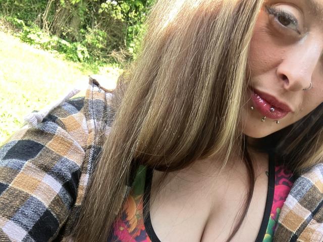 Harley Quinnzel, avec de longs cheveux bruns, une chemise à carreaux, un haut coloré et des lèvres roses avec des piercings, montrant sa poitrine et portant un petit anneau de nez, debout à l'extérieur sous une lumière du soleil vive..