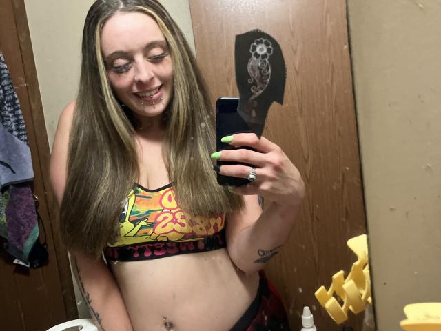 Harley Quinnzel met lang bruin haar, lachend, die een kleurrijke sportbeha draagt, haar borsten laat zien, een telefoon vasthoudt, neon groene nagels heeft, tatoeages en een neusring, in een badkamer..