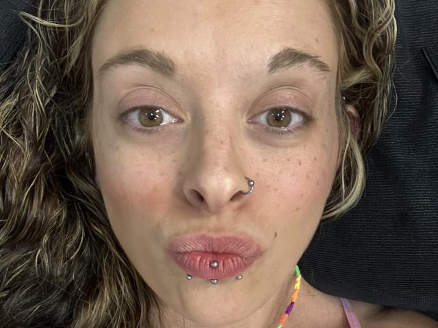 Harley Quinn heeft een neusring, een lipdoorboord met twee steentjes, en een kleurrijke ketting. Ze maakt een fronsende gezicht met bruine ogen en golvend blond haar..