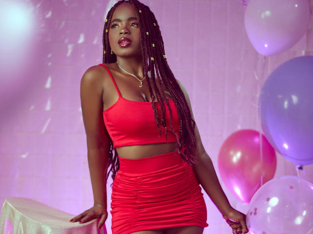 LindaBird with dark skin and long braids wearing a red crop top and ruched red skirt, standing with hands on a table, surrounded by colorful balloons.