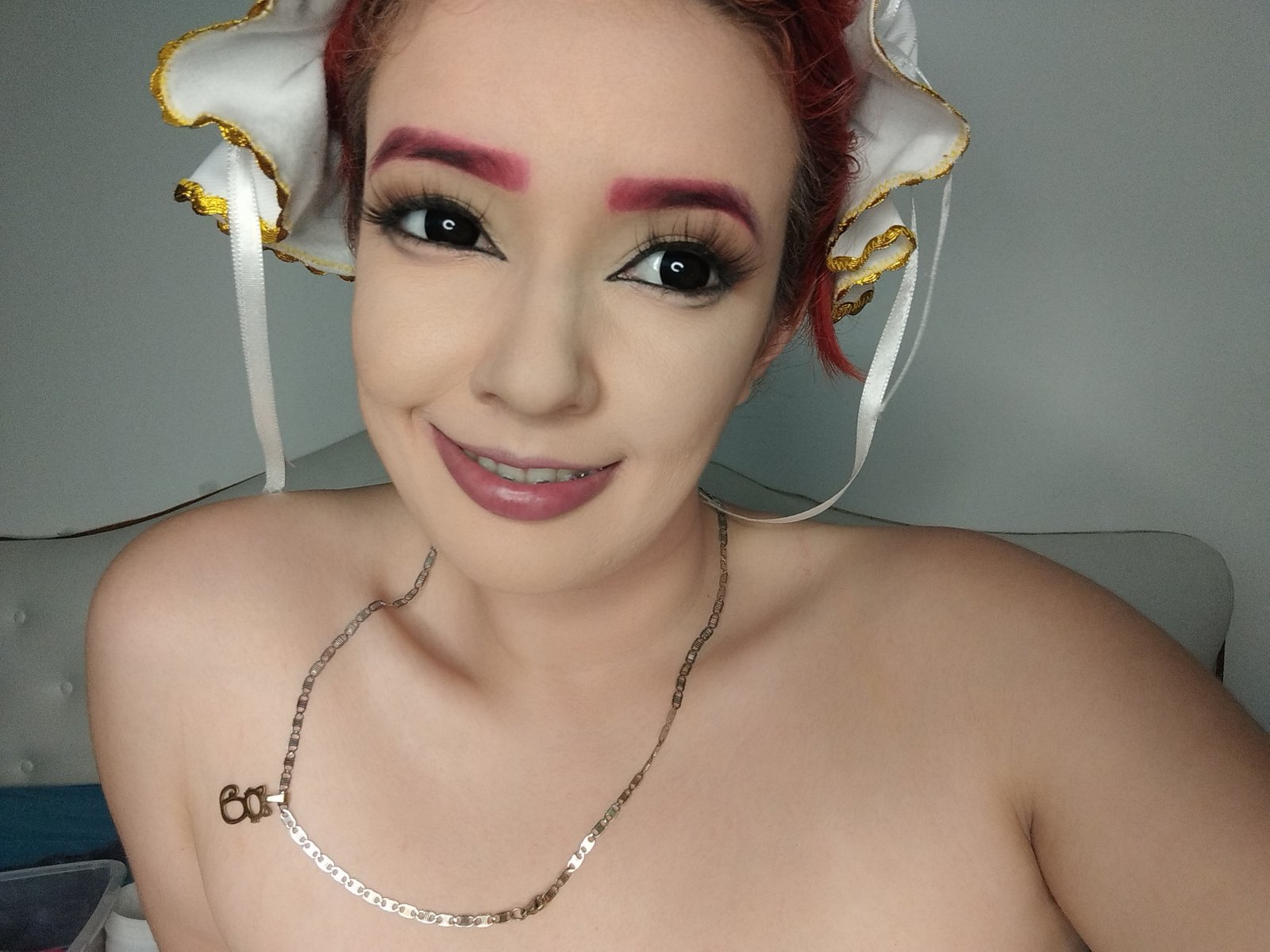 MadelineFoxy with red hair, white headpiece, pink eyebrows, black eyeliner, smiling, pink lipstick, gold necklace, bare shoulders, small tattoo on right side, and a cute nose piercing.