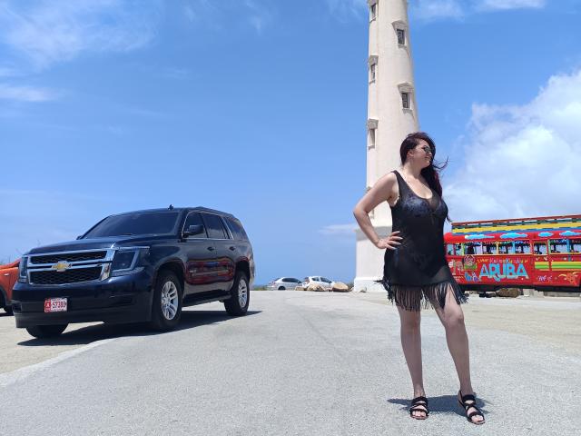 KatherinLatin stands in a black fringed dress, hands on hips, next to a black SUV, with a colorful bus and tall tower in the bright, blue sky.