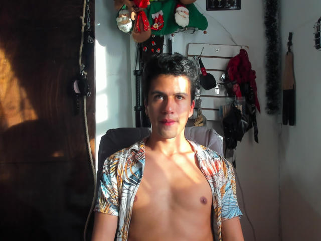 Shirtless DomStravelers with short black hair, light skin, and a slight smile, wearing a colorful floral shirt, sitting in a chair, sunlight on his chest, in a room.