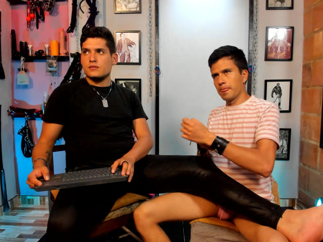Two muscular DomStravelers, one in black shirt and jeans, holding keyboard, other in pink striped shirt, black leather pants, raising foot, in dimly lit room, wearing wristband.