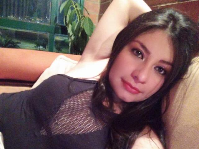 MaidenGame with long brown hair, pink lipstick, and black mesh top, lying on a couch, arms behind head, showing small tits, slightly smiling, in a cozy indoor setting.