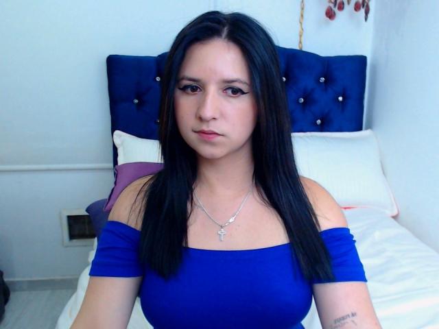 Kkatalina with long black hair, blue off-shoulder top, and silver necklace, has a serious expression and a tattoo on her right forearm, sitting on a bed.