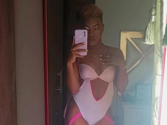 LilaJamez in a pink and white one-piece with a deep cleavage, holding a phone, showing off her tits and ass, with blonde hair in a bun.