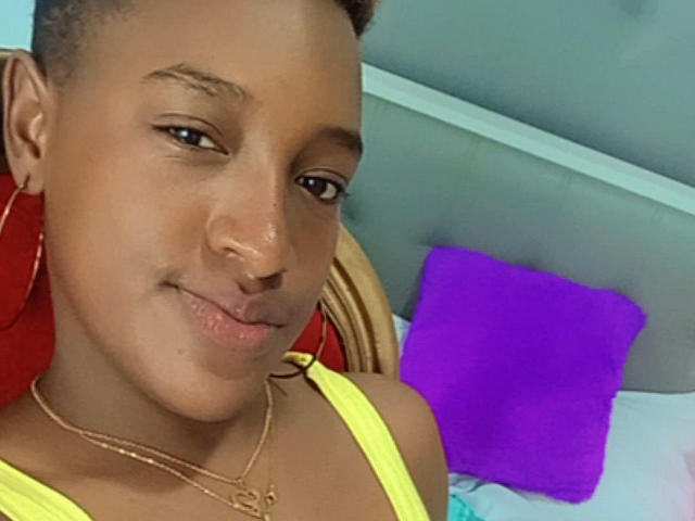 LilaJamez with short hair, brown skin, hoop earrings, gold necklace, yellow top, pouty lips, purple pillow in background, looking at camera with slight smile.
