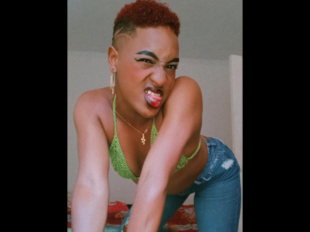 LilaJamez, with short red curly hair, green crochet bikini top, and blue jeans, leans forward, sticking out her tongue and making a playful, angry face. Her tits are small.