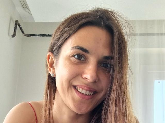 KieraHayes has light brown skin, straight brown hair, small diamond earrings, and is smiling with visible teeth, wearing a red spaghetti strap top, showing her nose and lips.