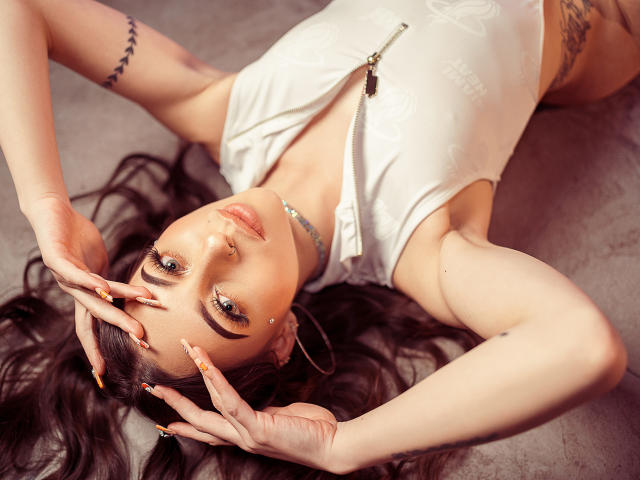 SamantaKnoxx lying on her back, hands on her head, wearing a white zip-up top, showing tattoos, with long dark hair, blue eyes, and a nose ring.