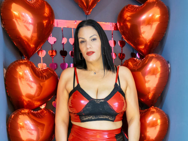 LizaRussell with black hair, red and black lace-up bra, showing her boobs, surrounded by shiny red heart balloons, blue wall, red heart strings, and a subtle necklace.