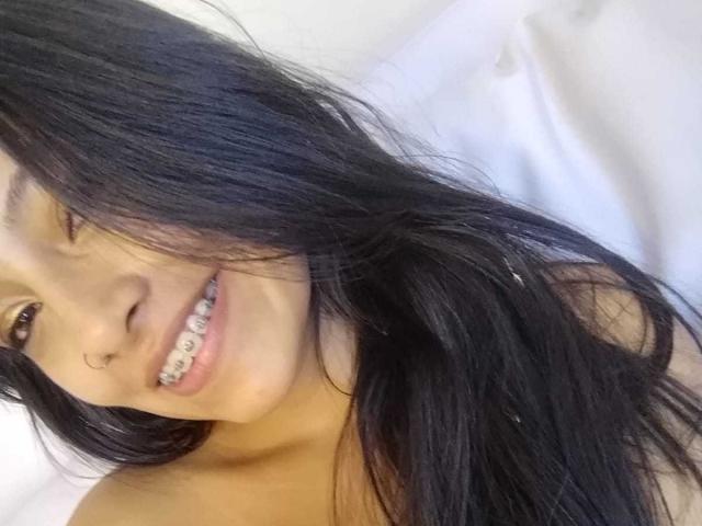 NoremCute with long black hair, smiling with teeth visible, showing metal braces, has a light brown skin tone, and is in a white-clothesed setting, looking cute and slightly goofy.