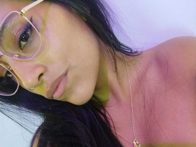 NoremCute has tan skin, long black hair, and is wearing gold-rimmed glasses with purple eyeshadow. She's looking at the camera with a slight pout, necklace visible.