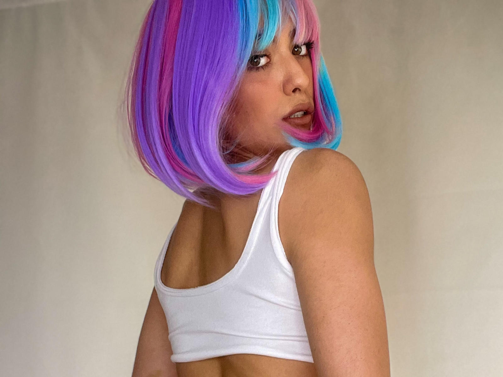 PortraitMarie with a colorful purple, pink, and blue bob wig, looking over her shoulder, wearing a white sports bra, showing her bare back and small, perky tits.