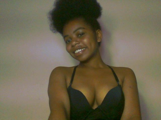 RayOfBeauty has dark skin, a cute afro, and is smiling at the camera. She's wearing a black bra that highlights her tits. The photo is slightly blurry.