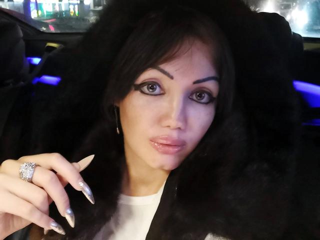 SexyNat-hot with shiny black hair, heavy eyeliner, glossy pink lips, long silver nails, and a fur coat, showing a ring, in a car at night.