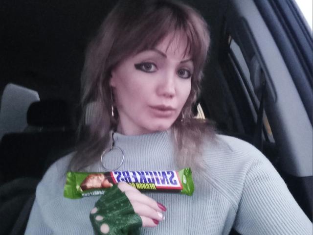 SexyNat-hot with brown hair, wearing a gray sweater, holding a green and yellow candy bar in her right hand, and a ring in her left ear, sitting in a car.