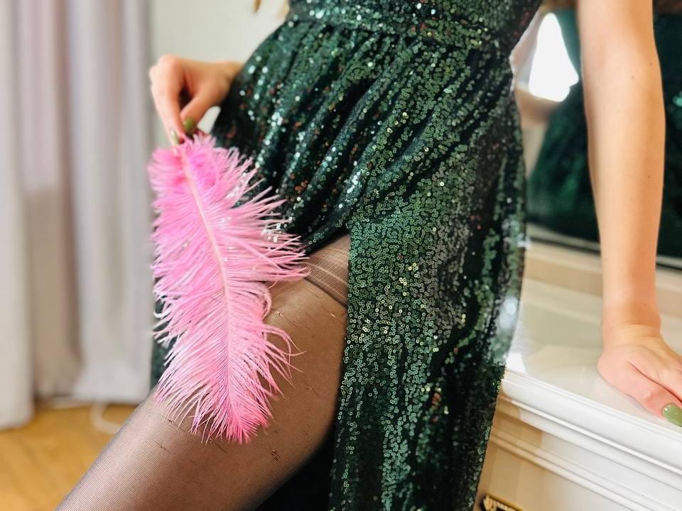 LeylaLo's sequined green dress, sheer black stockings, holding a pink feather against her thigh, hand resting on a mirror, wooden floor, green nail polish, sexy, flirty.
