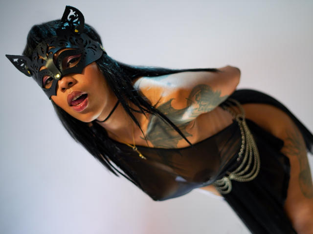 InannaIsshtar with black cat mask, black sheer dress, and rope, leaning forward, mouth open, tattooed arms, long black hair, visible boobs, and tattooed lower body, looking sexy.
