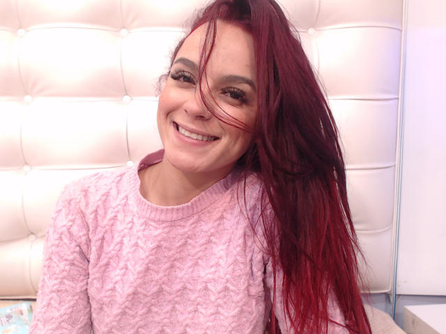 MaiaPark, com longos cabelos ruivos, sorrindo, vestindo um suéter rosa, texturizado e fofo. Seus seios são pequenos e suas nádegas não estão visíveis. Ela está sentada em frente a uma parede branca, com textura..