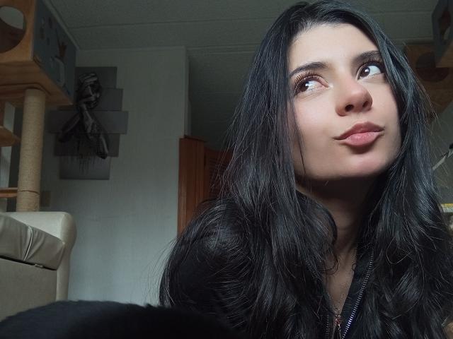 JeneKurz with long black hair, looking up with slightly pouty lips, wearing a black top, in a dimly lit room. Her brown eyes are gazing upwards.