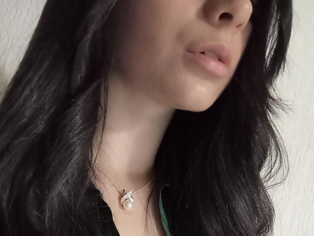 JeneKurz's face with black wavy hair, pale skin, slightly parted pink lips, and a silver necklace with a pink gem. Boobs not visible, focus on upper chest.