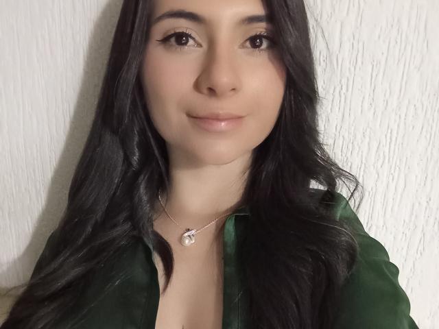 JeneKurz has long black hair, a light skin tone, and is wearing a green shirt with a silver necklace, smiling softly at the camera. Boobs slightly visible.