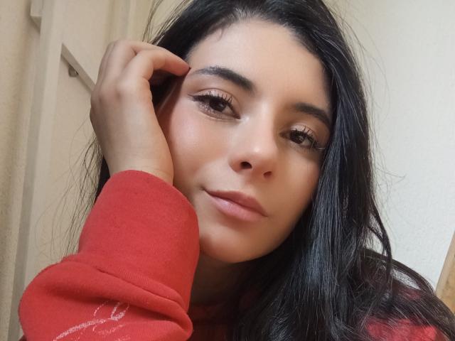 JeneKurz has long black hair, wearing a red sweater, with a hand on her cheek, brown eyes, and full pink lips, looking slightly to the right.